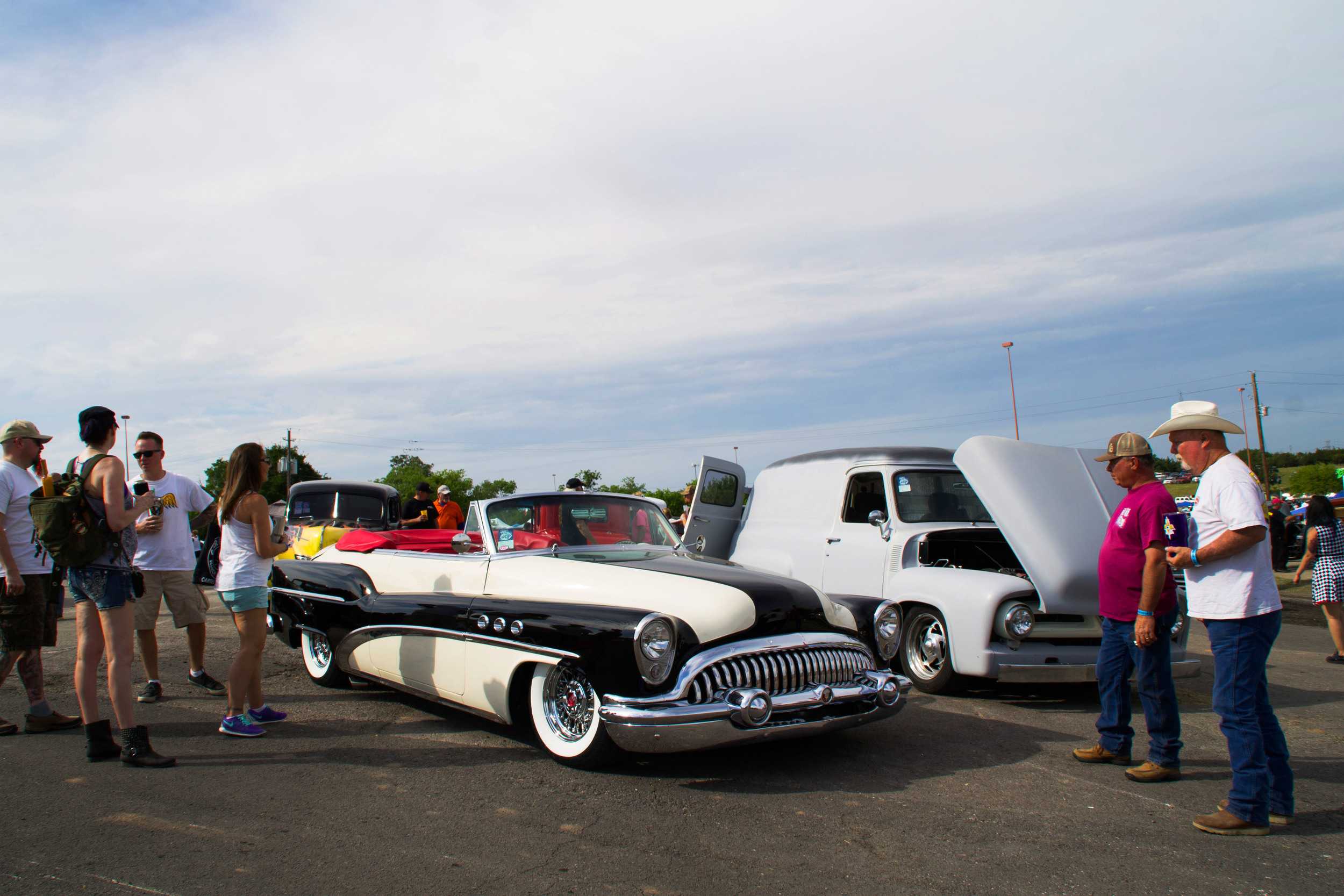  People stand around, scrutinizing a glossy black and white vehicle. People of all ages joined together to enjoy the wide variety of cars present. 