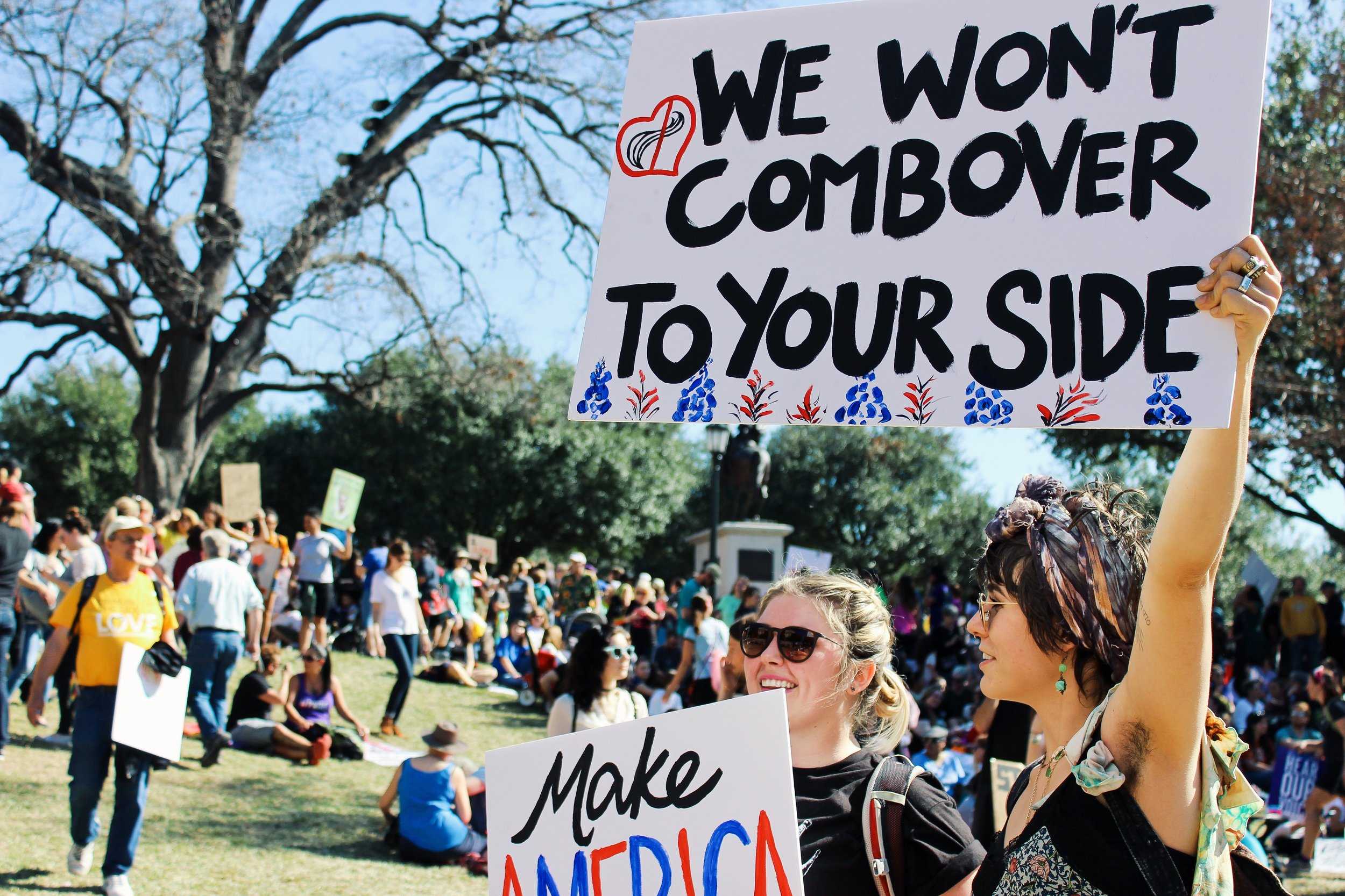   Two women stand with their signs at the march. One sign says, "We won't combover to your side."  