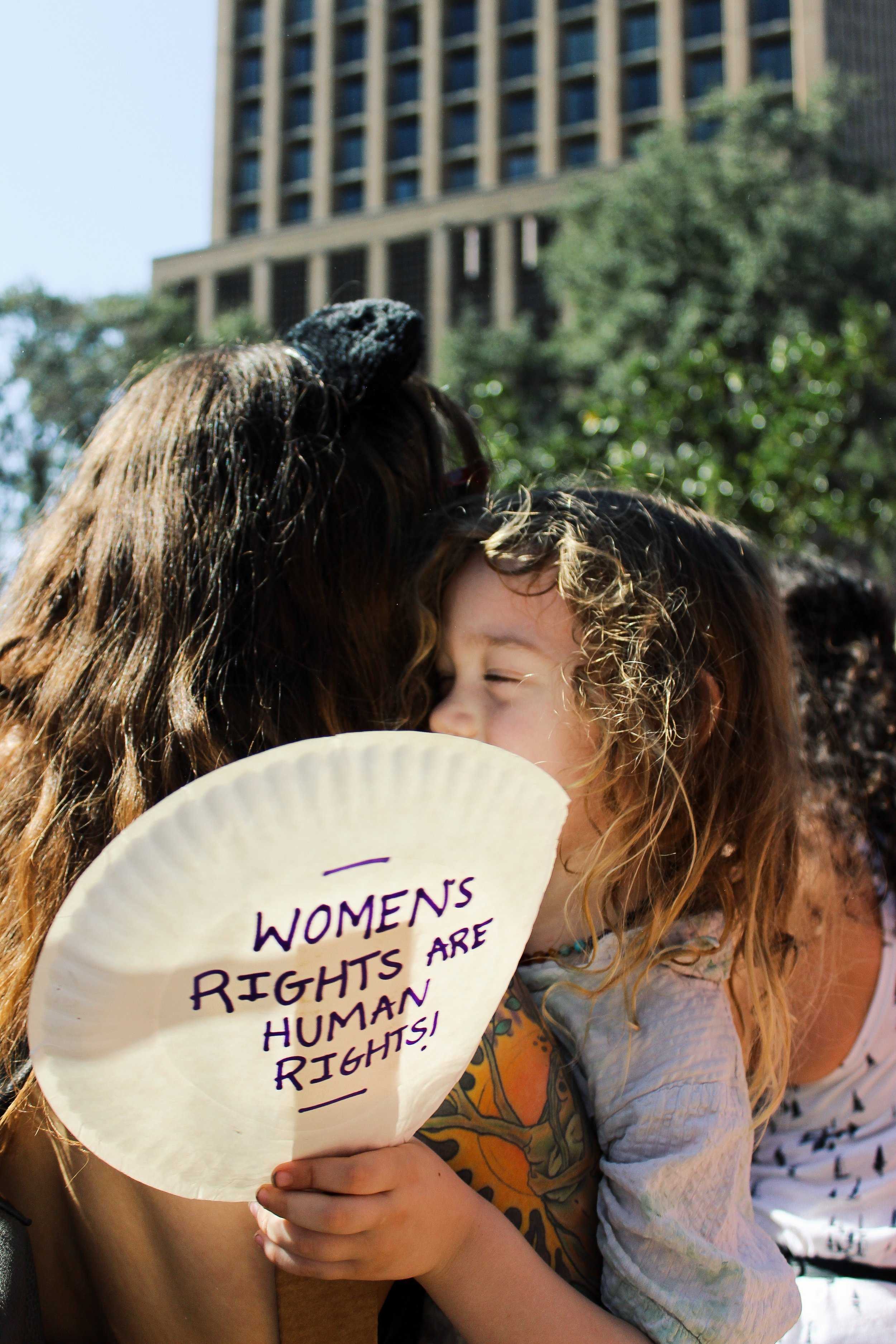   A young girl hugs her mom at the march, holding a handmade sign that says, "Women's rights are human rights!"  