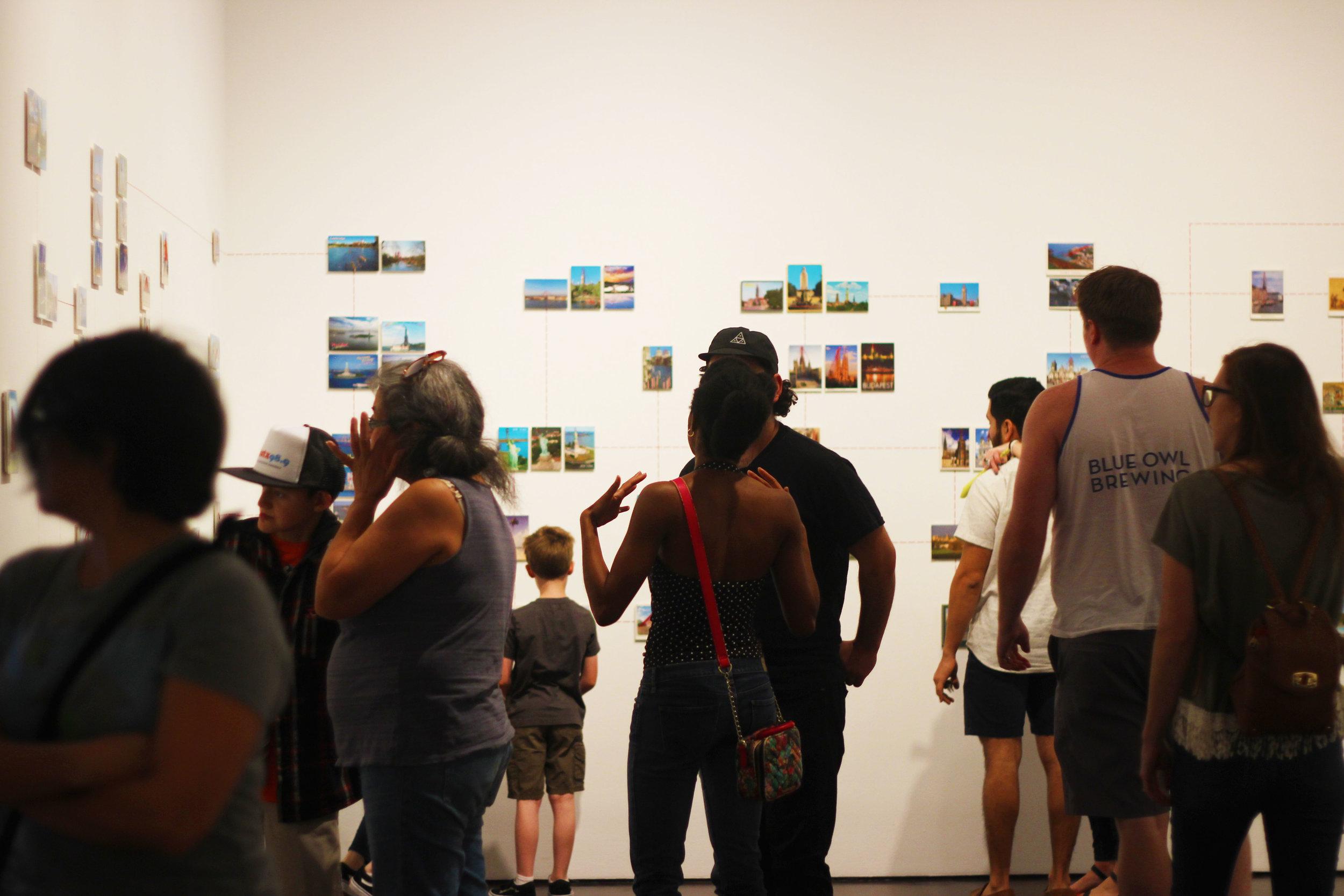   People walk through walls of “Paranormal Postcards,”&nbsp;a series a part of of the newly installed &nbsp;“Curiouser” exhibit that interconnects a world view through postcards and red thread.  