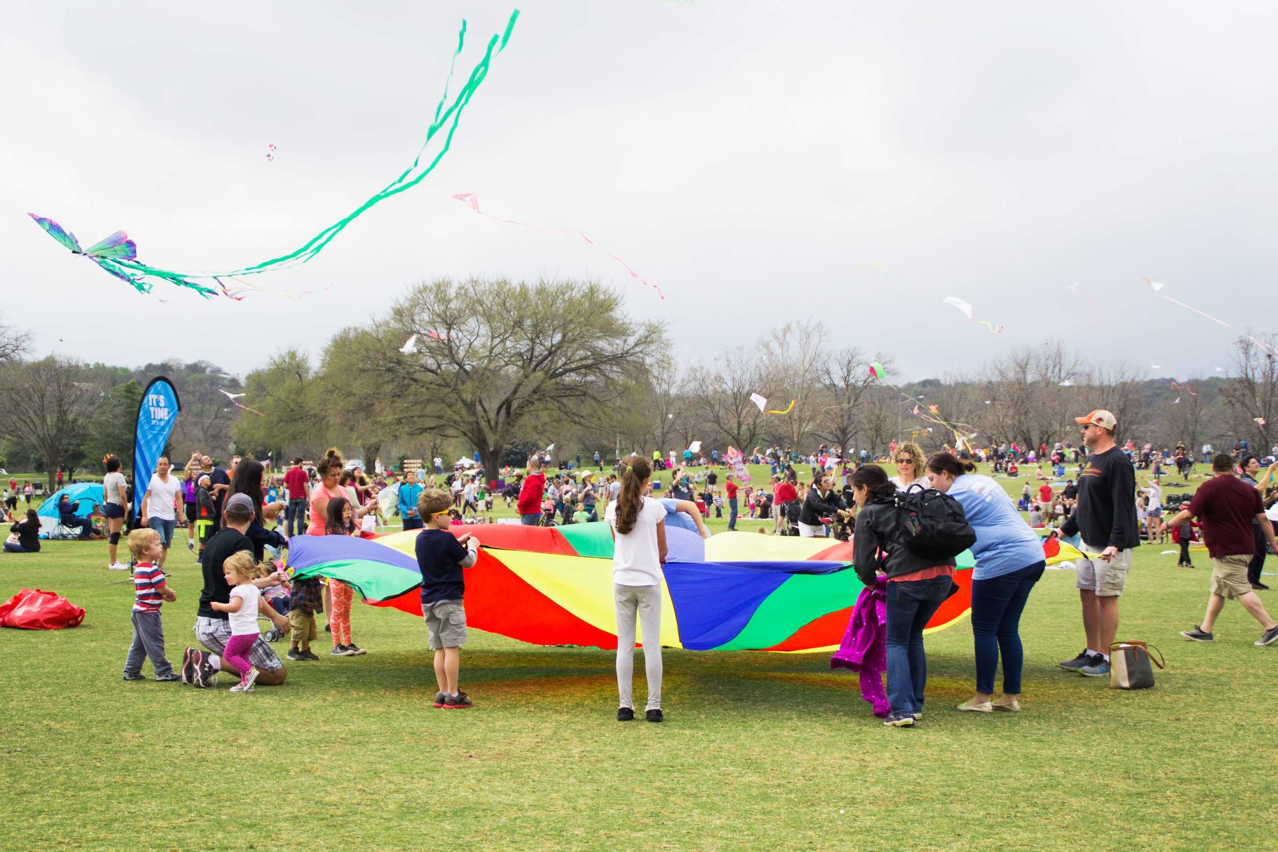  People bounce a ball on a colorful parachute while kites soar overhead. 