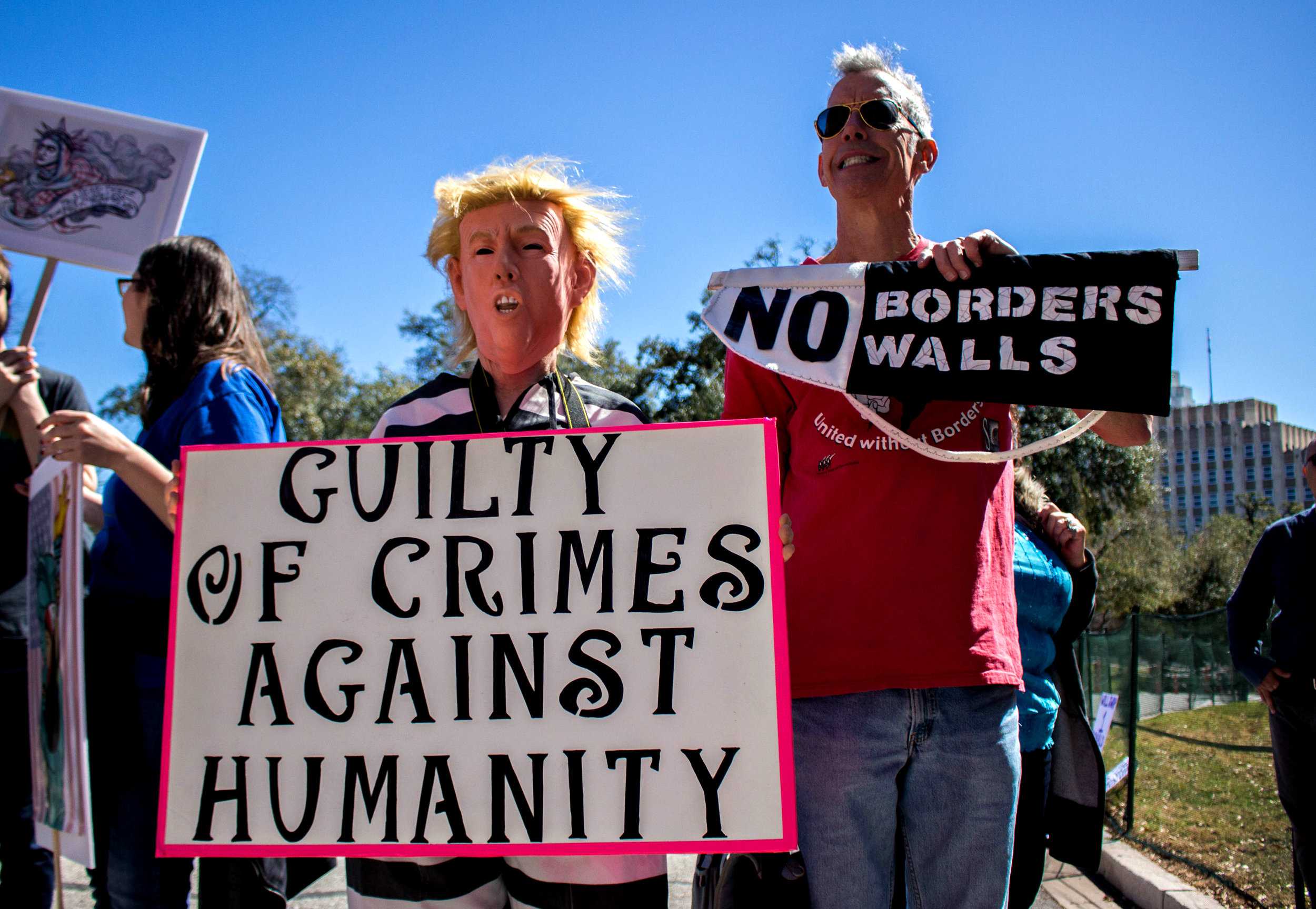   Two protestors hold a signs that say, "Guilty of crimes against humanity" and "No borders/walls" at the rally. One protestor is wearing a Trump mask.&nbsp;  