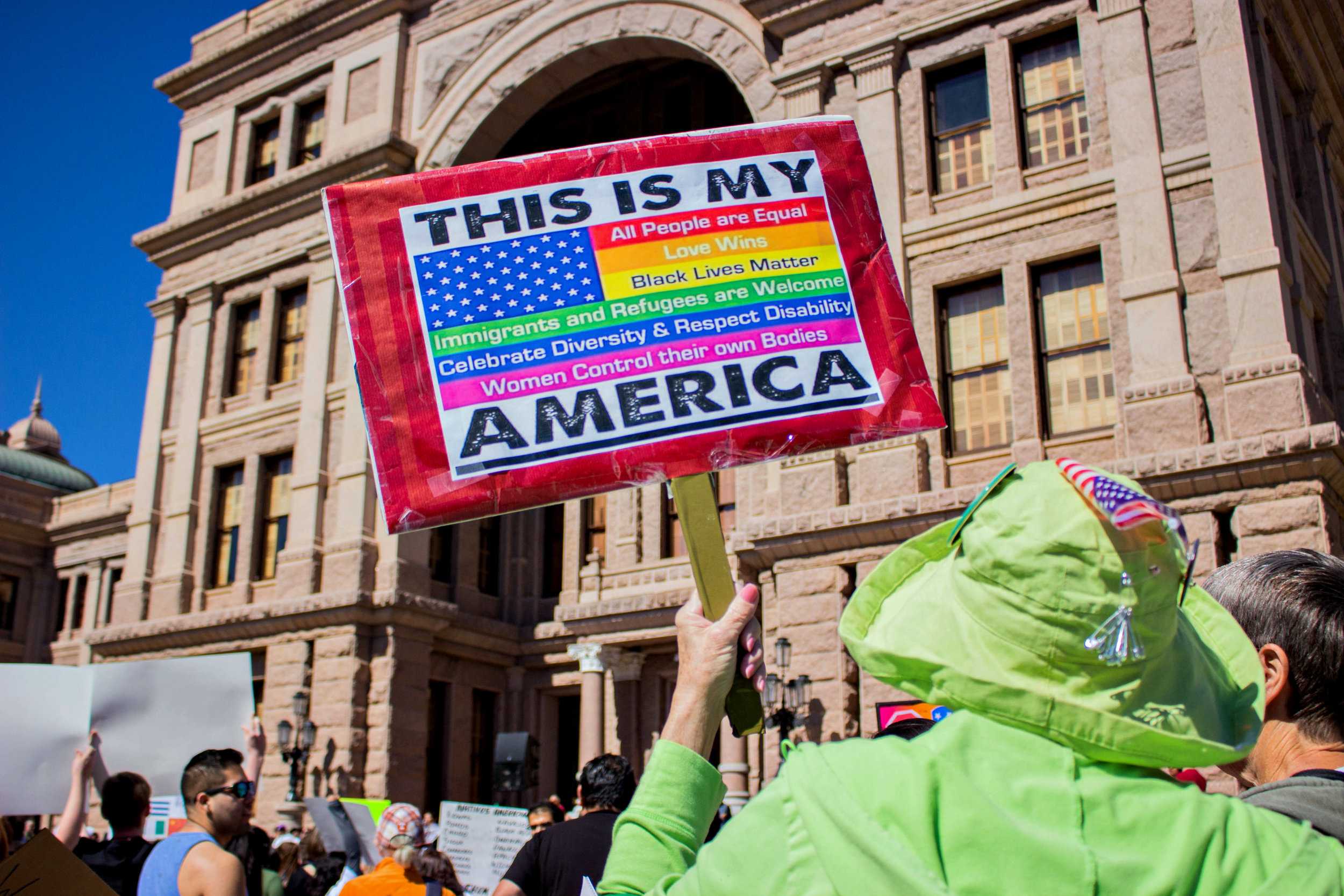   A protestor holds a sign that says, "This is my America" with a rainbow American flag.  